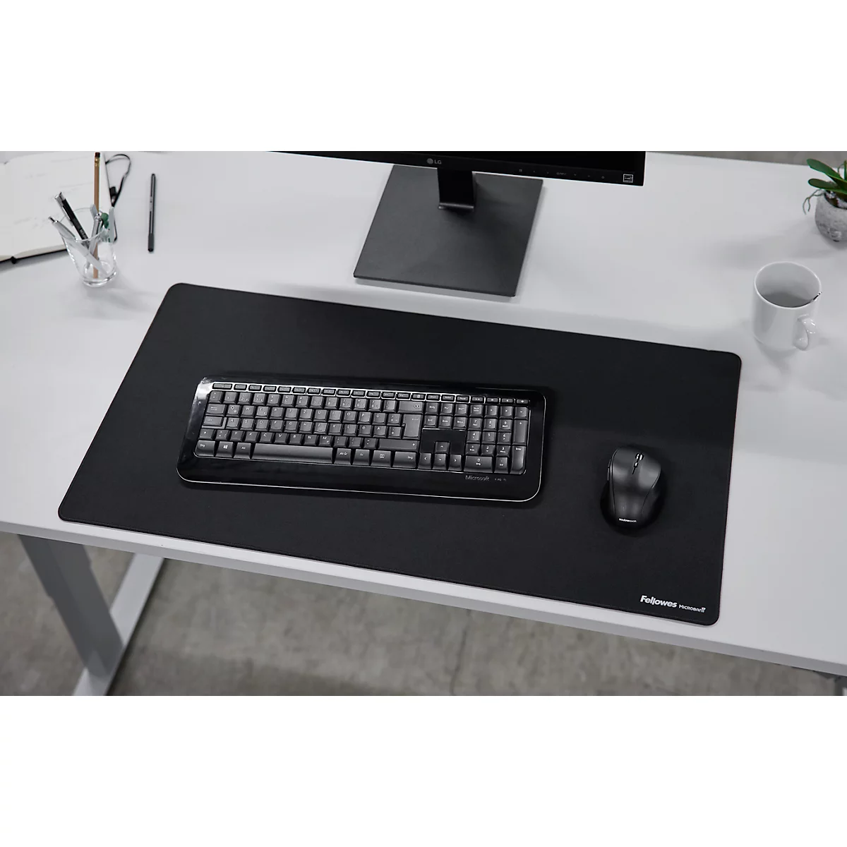 Schwarze Schreibtischunterlage mit Tastatur, Maus, Monitor und Becher auf weißem Schreibtisch.