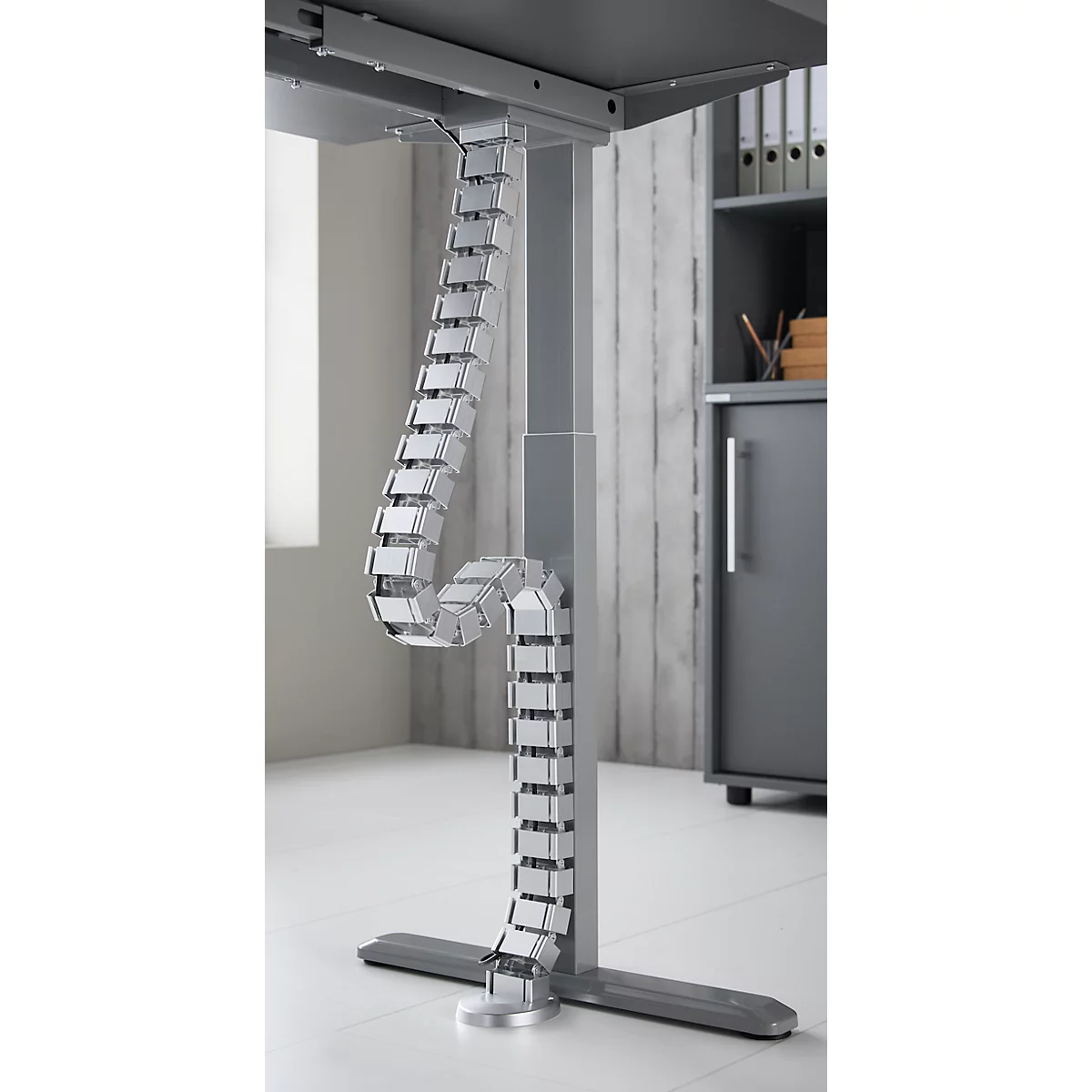 Kabelführung an einem höhenverstellbaren Schreibtisch, aus grauem Kunststoff. Der Schreibtisch steht im Büro.