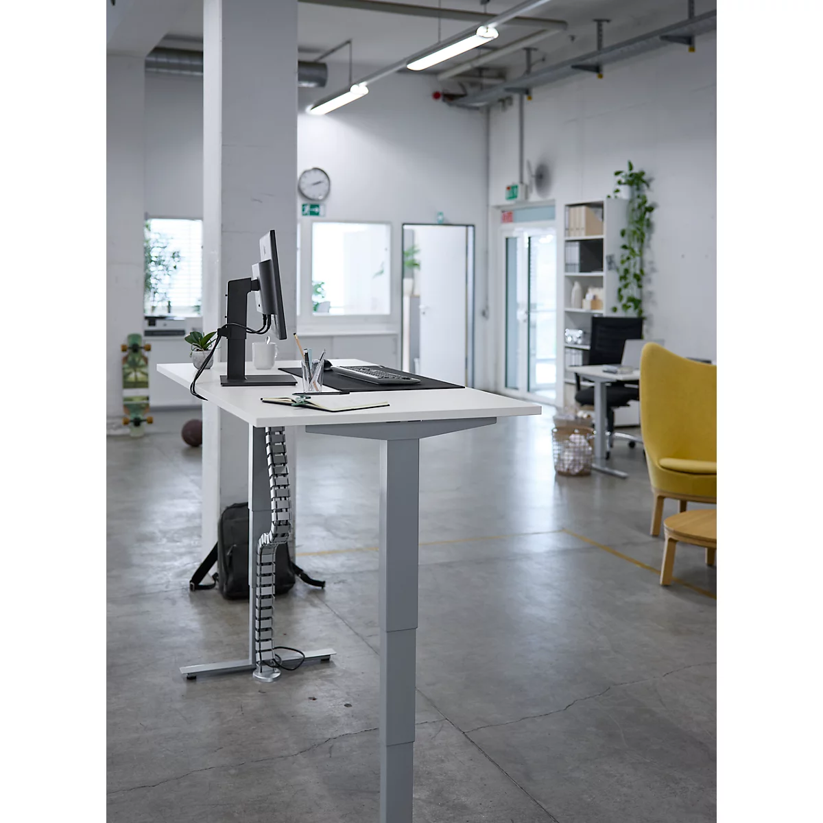 Ein höhenverstellbarer Schreibtisch in hellem Büro. Darauf Monitor, Tastatur und Kabel. Im Hintergrund ein gelber Sessel.
