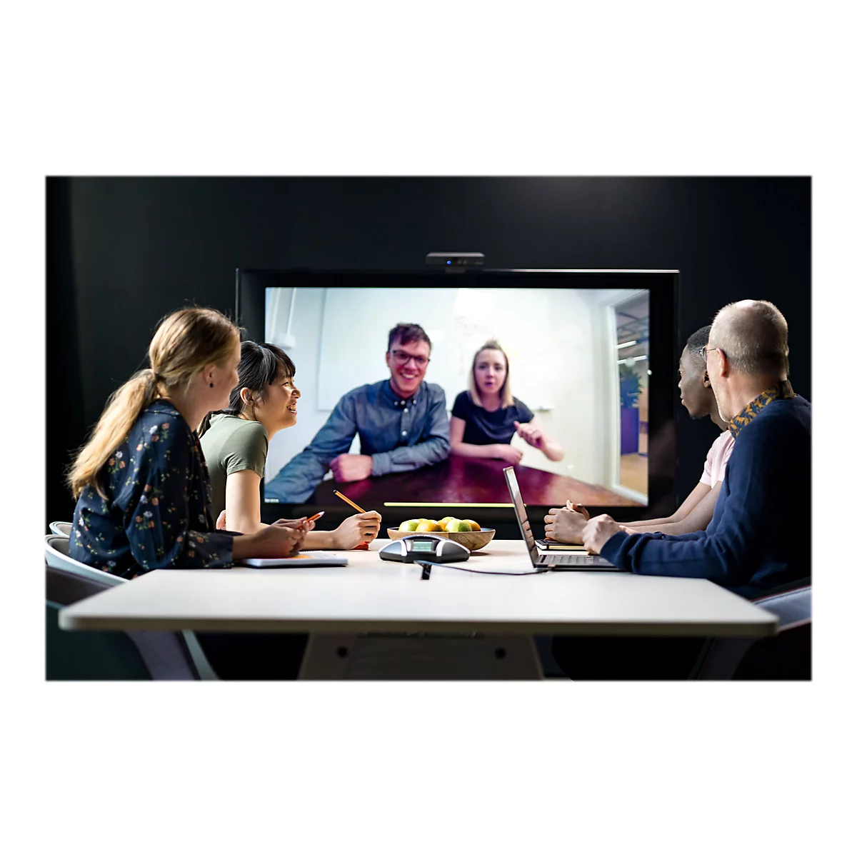 Gruppe von Kollegen in Videokonferenz. Auf dem Bildschirm: zwei Personen lachen. Auf dem Tisch: Obst, Laptop, Telefon, Notizbuch.