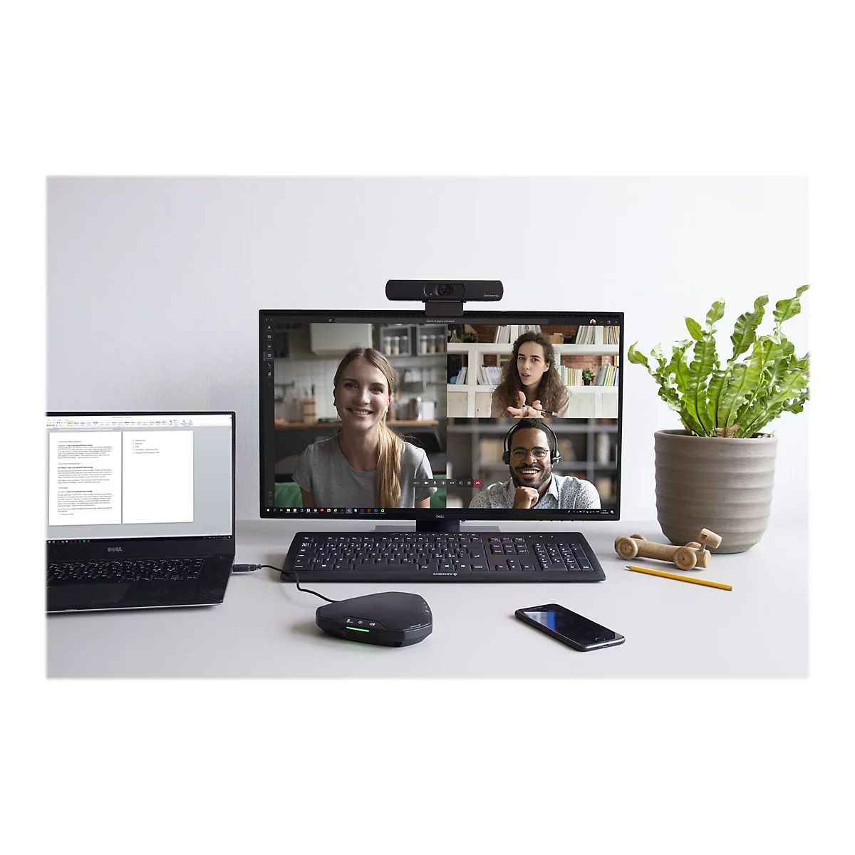 Eine Arbeitsplatzszene mit einem Monitor, der Videokonferenzteilnehmer zeigt. Darunter liegen Tastatur, Maus, Handy und Laptop.