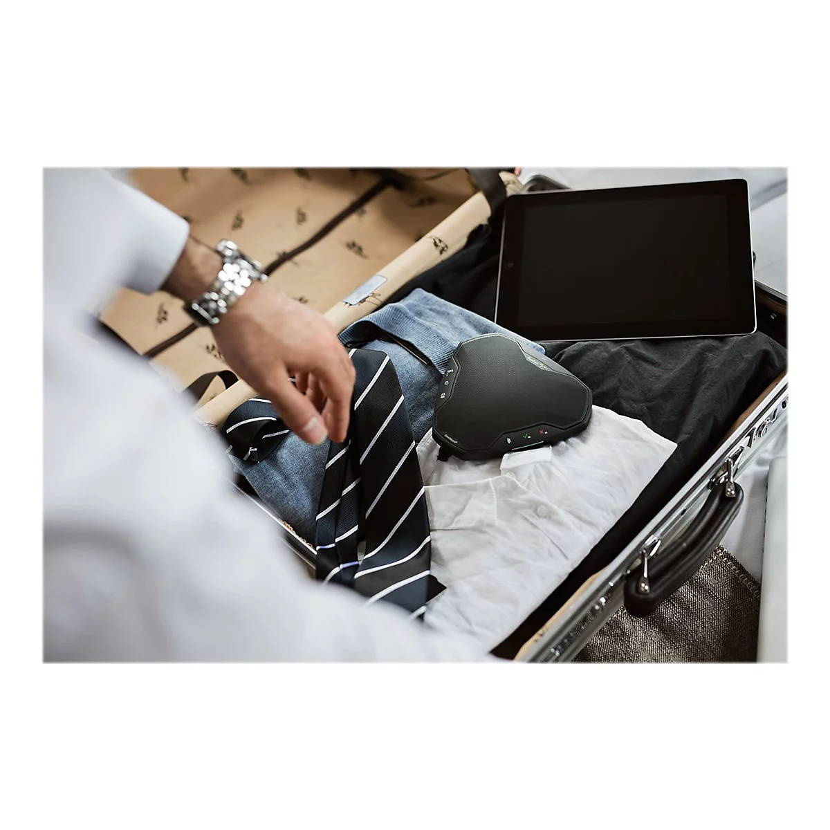 Ein Mann packt einen Koffer. Ein schwarzer ergonomischer Laptop-StÃ¤nder liegt zwischen Kleidung und einem Tablet.