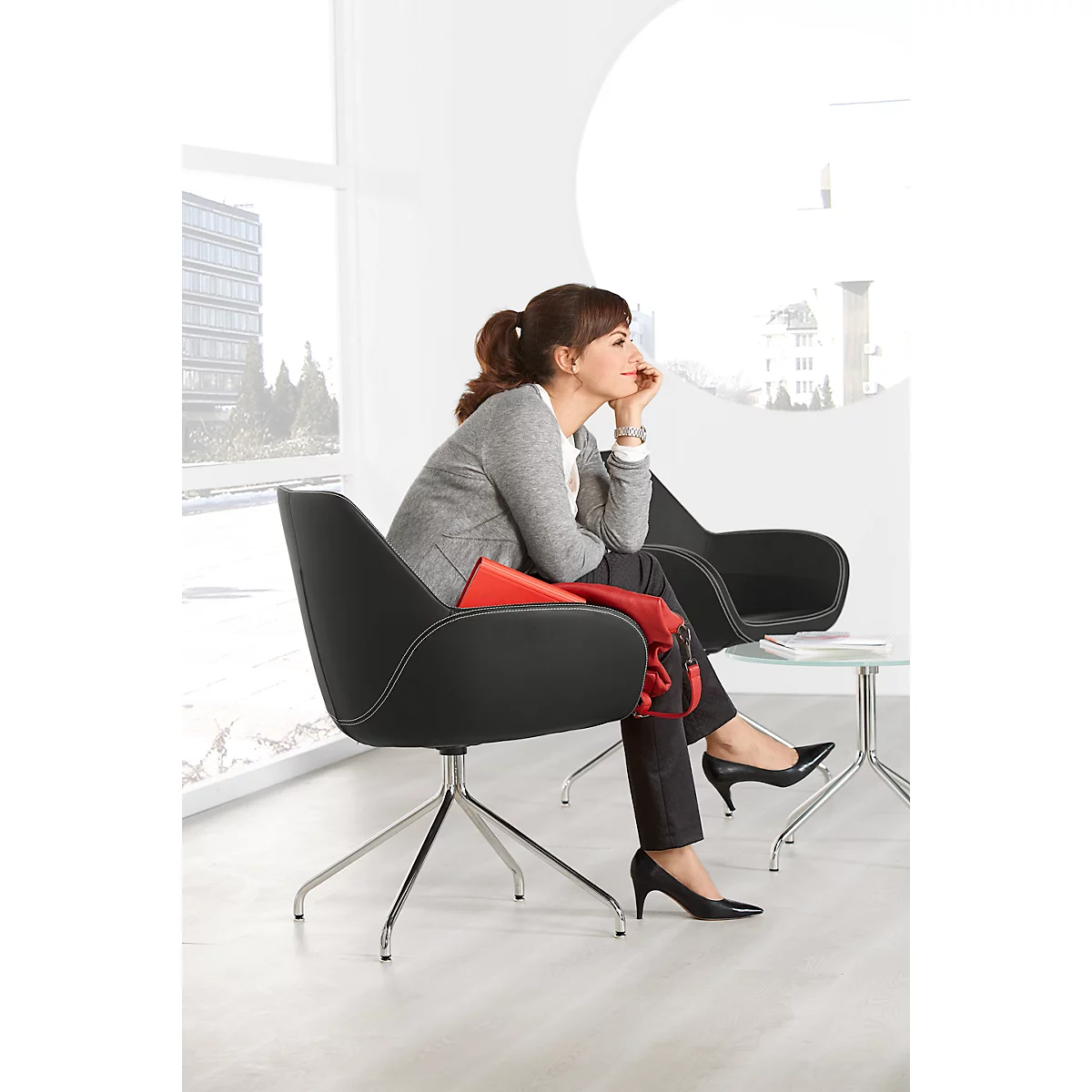 A woman in a gray blazer sits in a black chair, thoughtfully looking out the window.