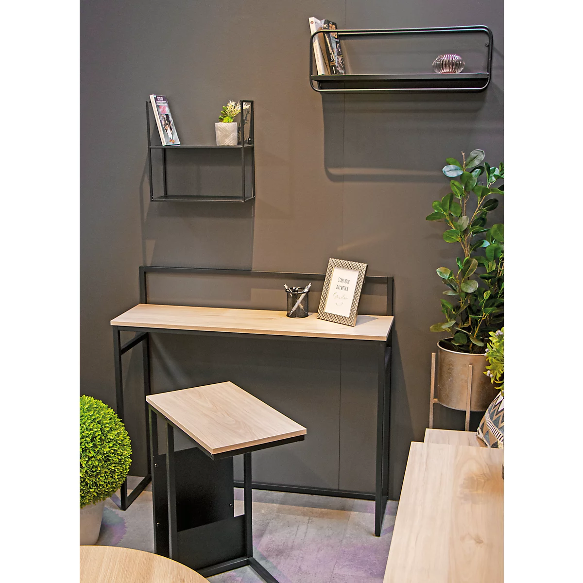 Schreibtisch mit Regal und Beistelltisch. Schwarzes Metallgestell, helle Holzplatten. Auf dem Schreibtisch Bilderrahmen und Stifte. Pflanzen als Dekoration.