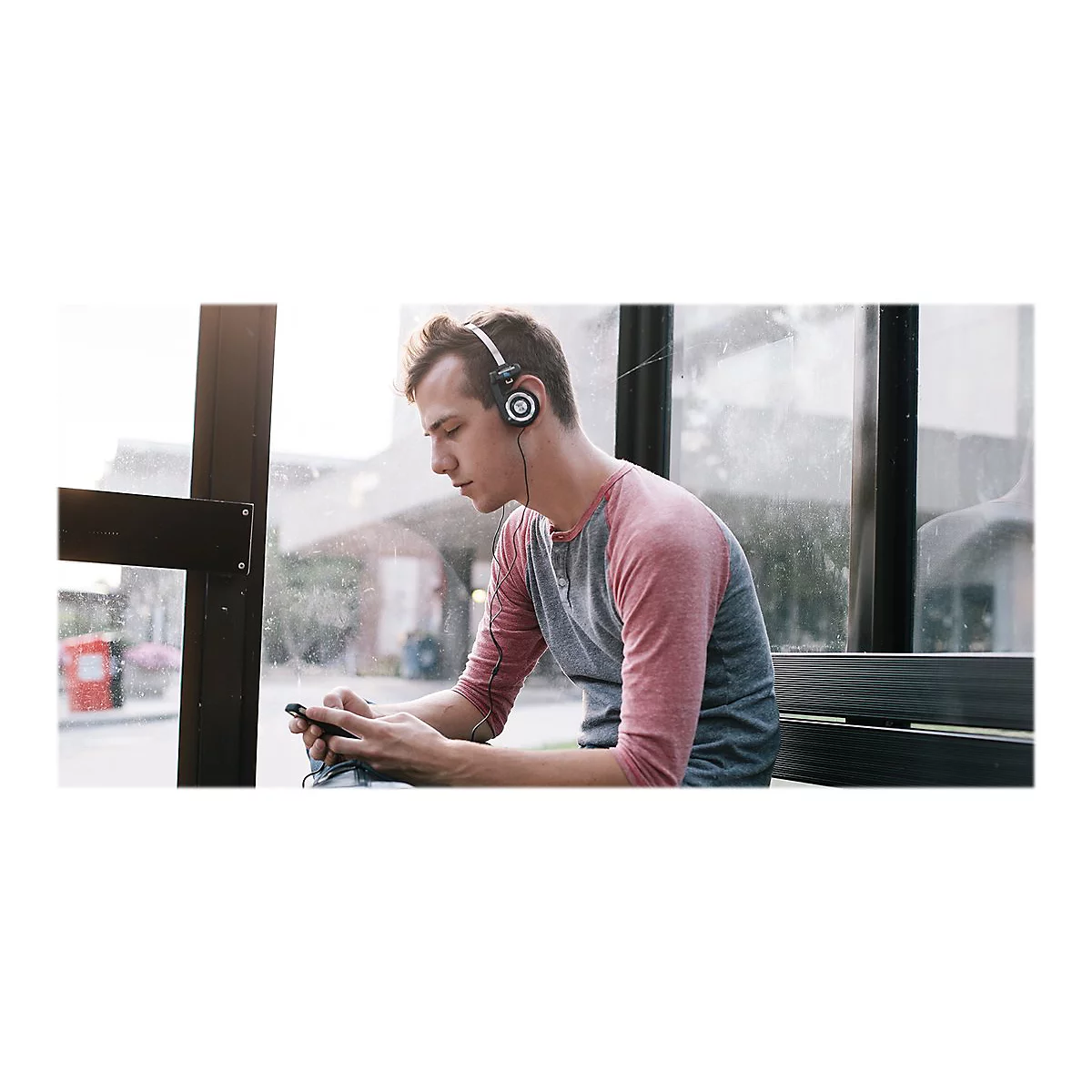 Ein junger Mann mit KopfhÃ¶rern sitzt in einer Bushaltestelle. Er blickt auf sein Handy, wahrscheinlich Musik hÃ¶rend.