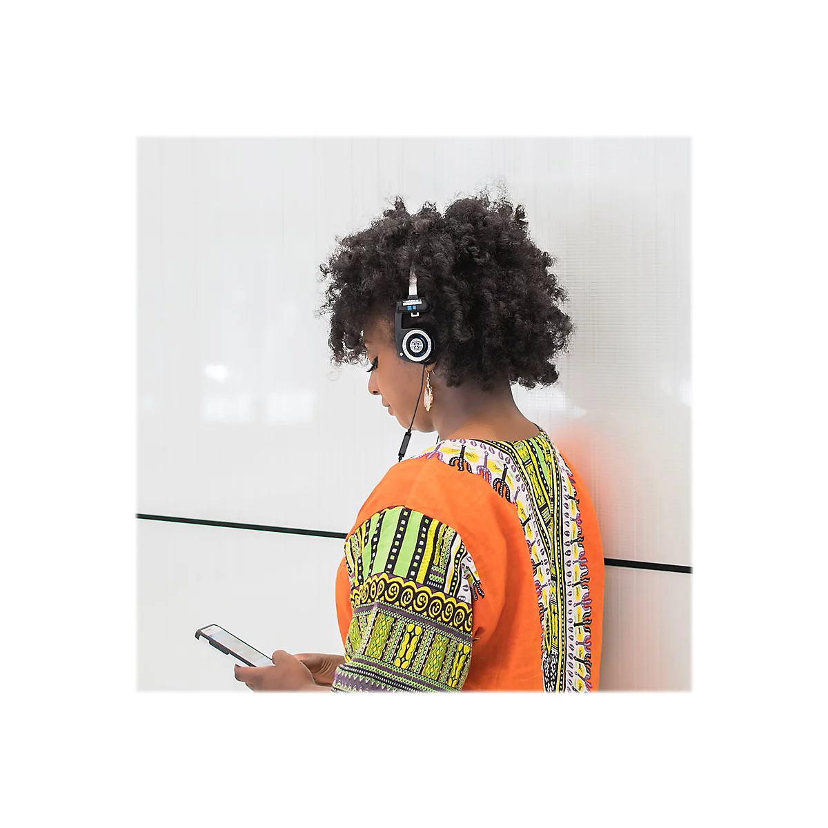 Eine Person mit Locken trÃ¤gt KopfhÃ¶rer, schaut auf ein Smartphone. Sie lehnt an einer weiÃŸen Wand, trÃ¤gt ein buntes Kleid.
