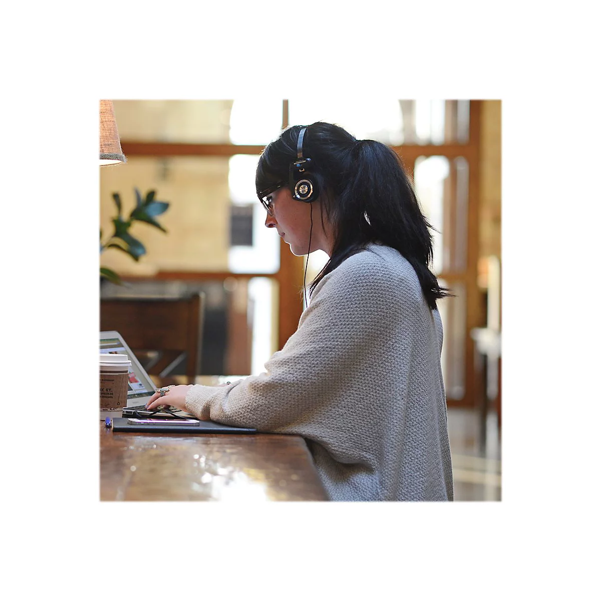 Eine Frau mit KopfhÃ¶rern und Brille arbeitet an einem Laptop. Sie sitzt an einem Holztisch.