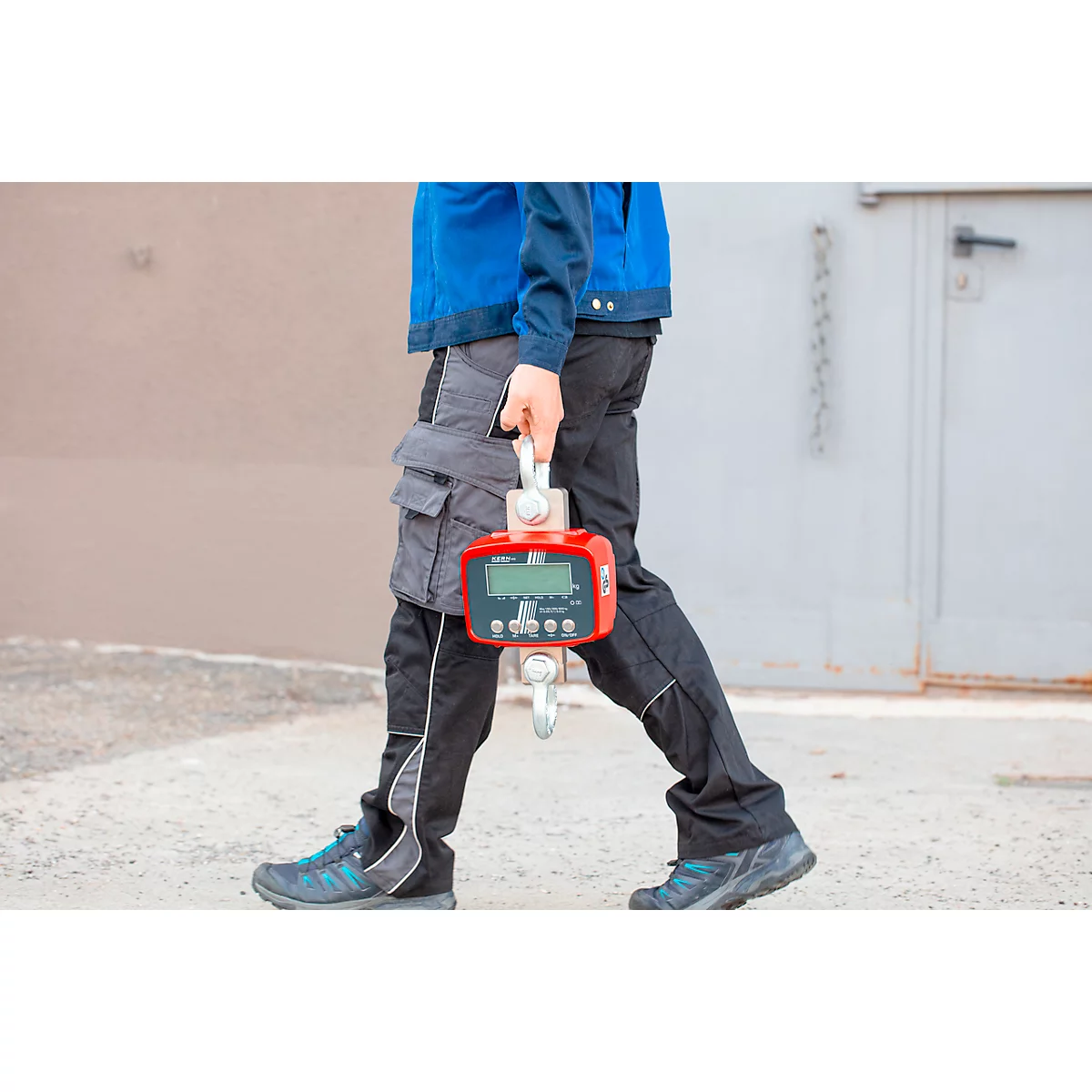 Eine Person trägt eine rote Waage mit Haken. Sie trägt eine blaue Jacke, schwarze Hose und geht an einer hellgrauen Wand vorbei.