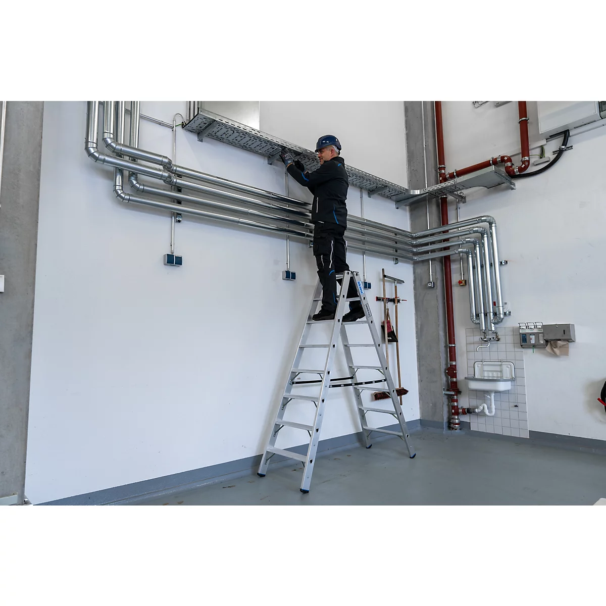 Een man staat op een ladder en werkt aan pijpen op een muur. Op de achtergrond: meer pijpen, een wastafel en gereedschap.