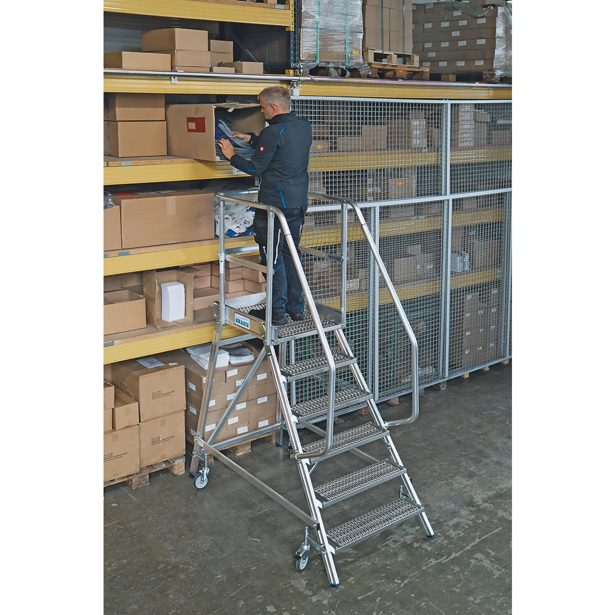 Een man staat op een trap en pakt spullen van een plank in een magazijn. De planken zijn gevuld met dozen.