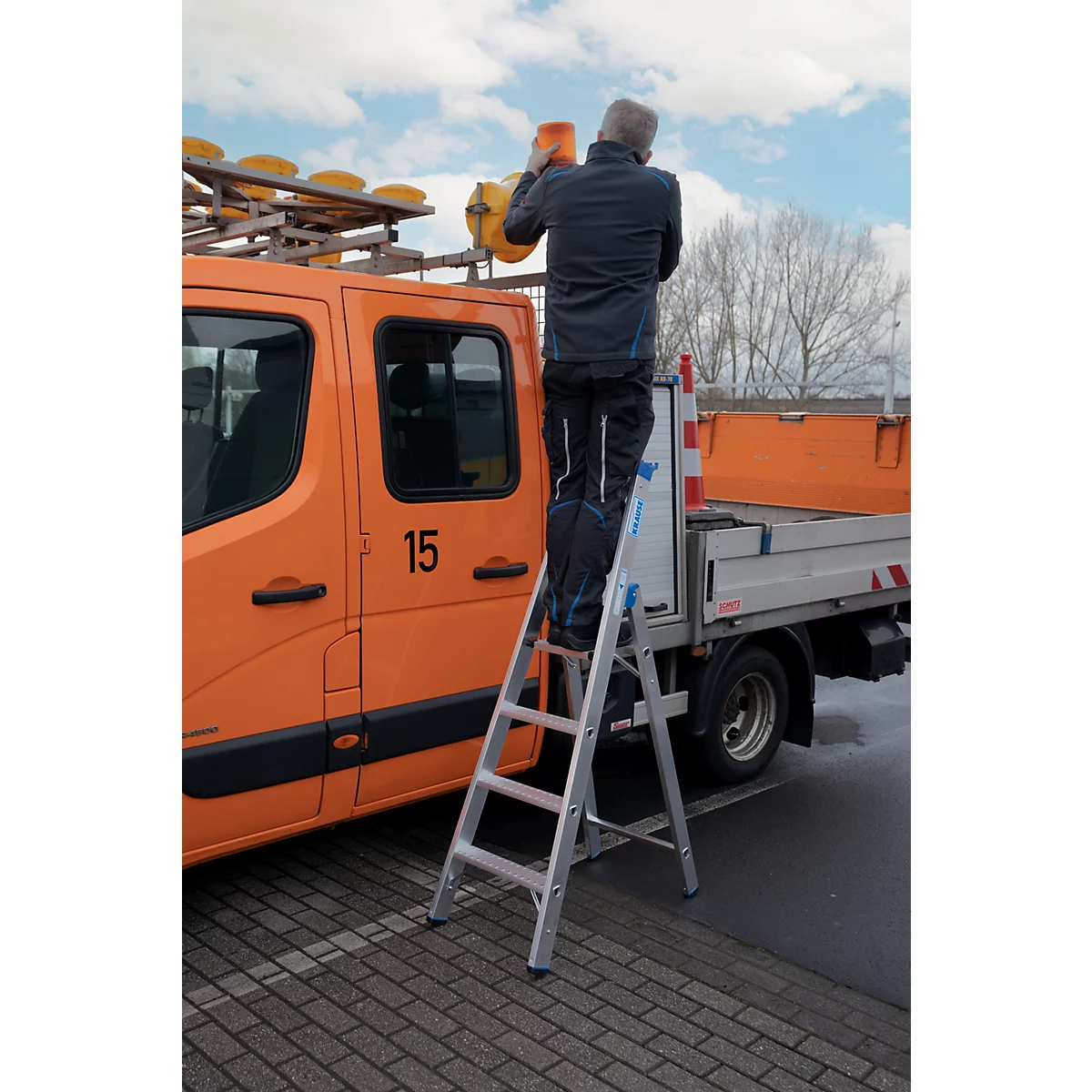 Ein Mann auf einer Leiter arbeitet an Warnleuchten eines orangefarbenen LKW. Im Hintergrund ein weißes Fahrzeug mit Warnmarkierungen.