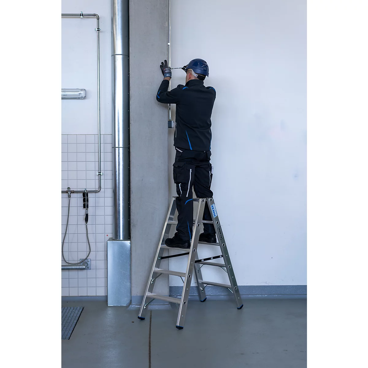 Ein Mann steht auf einer Trittleiter und schraubt etwas an einer Wand fest. Er trägt Arbeitskleidung.