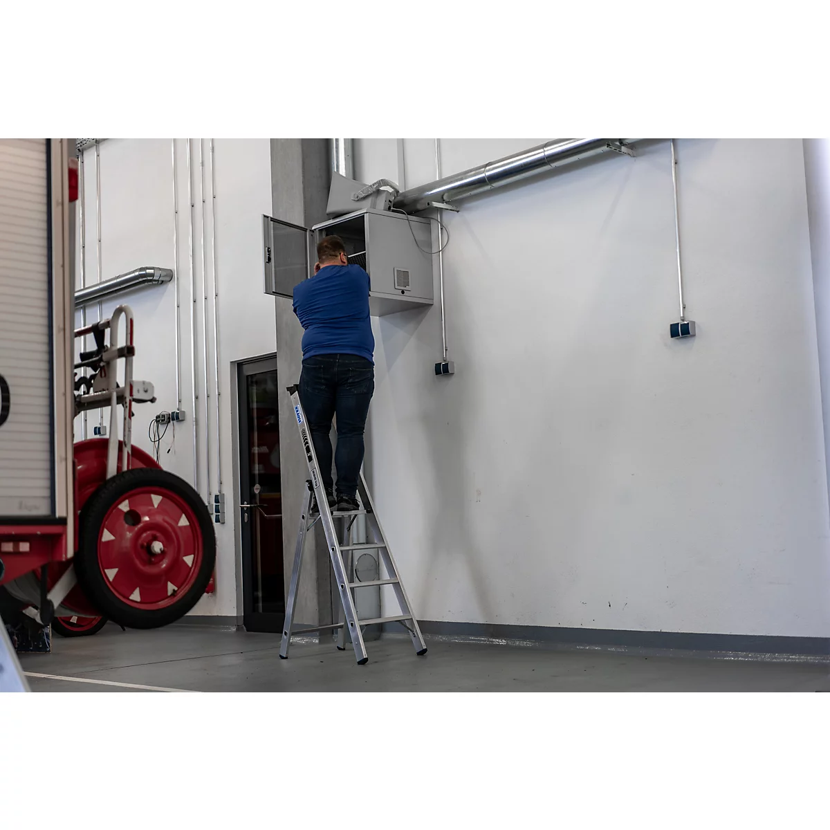 Een man staat op een ladder en werkt aan een open elektrische kast aan de muur. Naast hem is een brandweerwagen te zien.