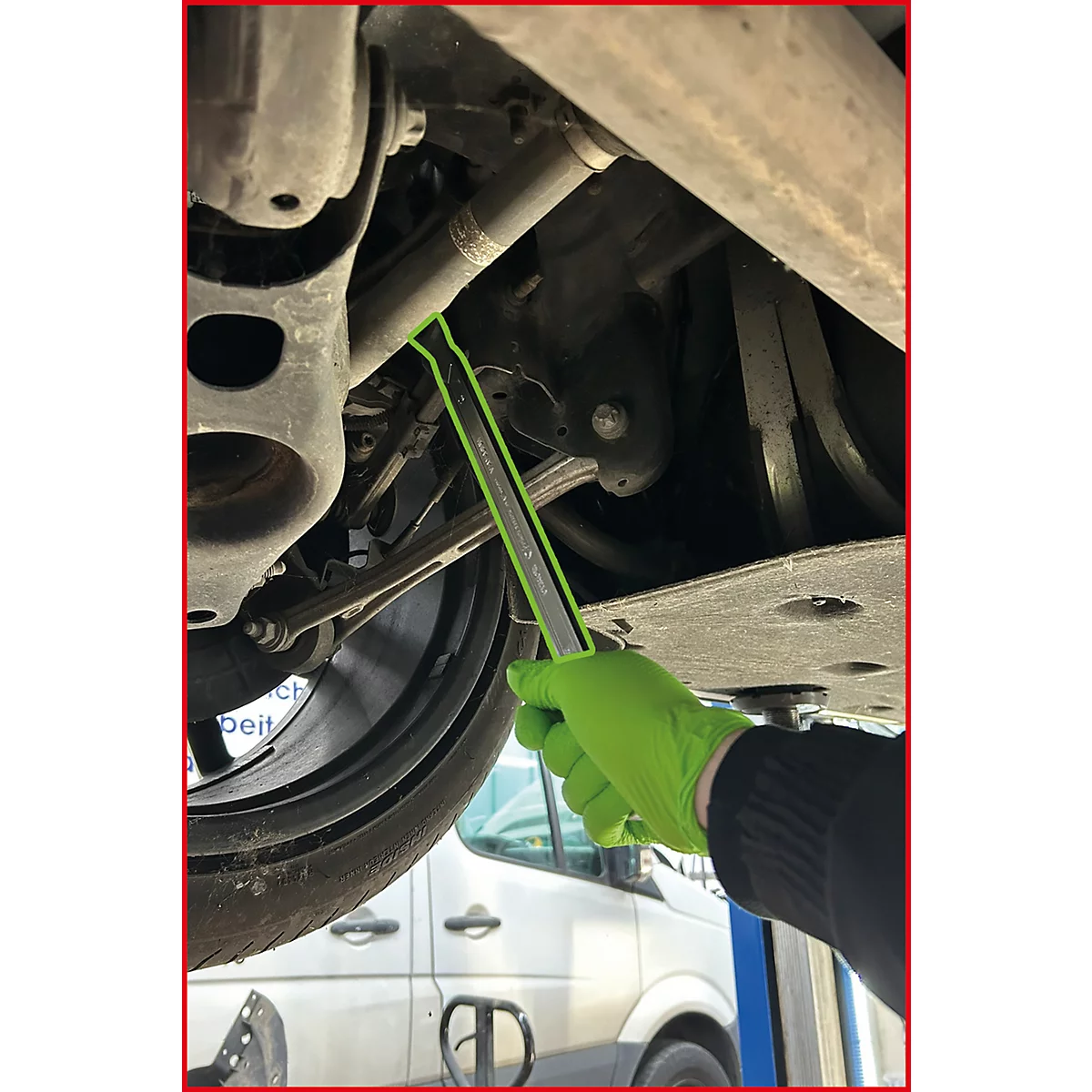 Hand mit grünem Handschuh benutzt ein Montiereisen an einer Autovorderachse in einer Werkstatt.