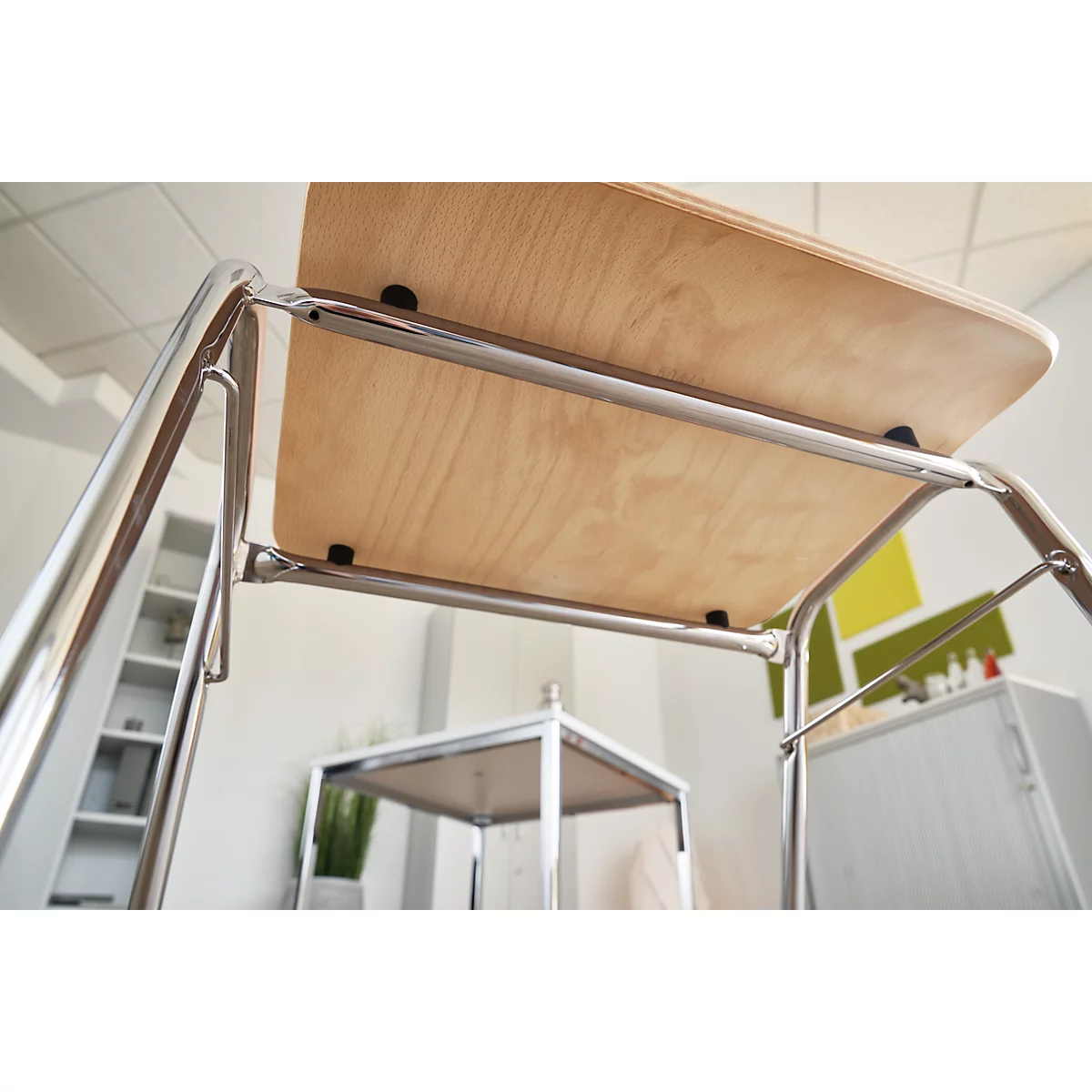 Houten tafel van onderaf gefotografeerd, met een verchroomd frame. Andere meubels en een plant op de achtergrond.