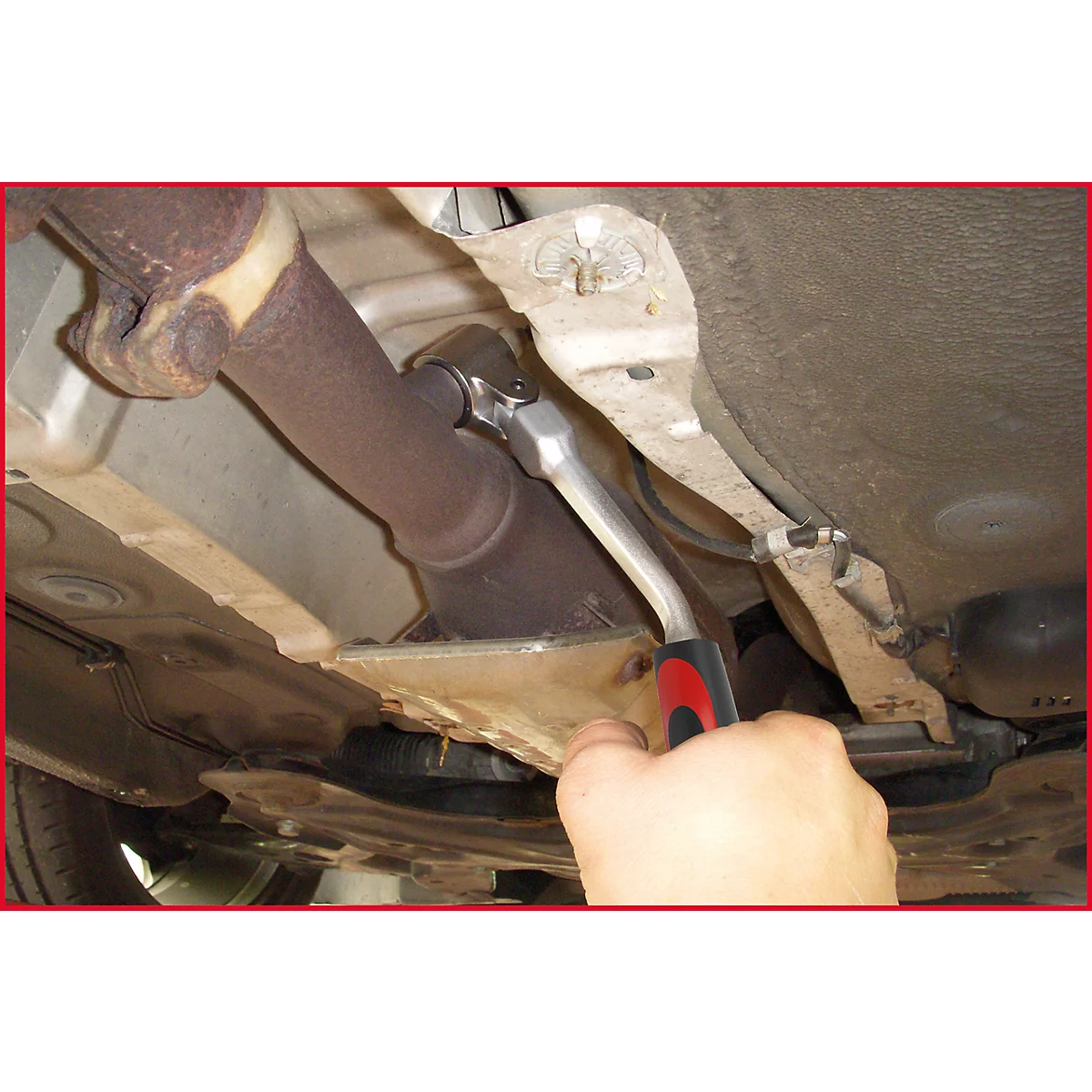 Ein Mechaniker löst eine Schraube an einem Auspuffrohr mit einer Ratsche. Die untere Fahrzeugseite ist zu sehen.