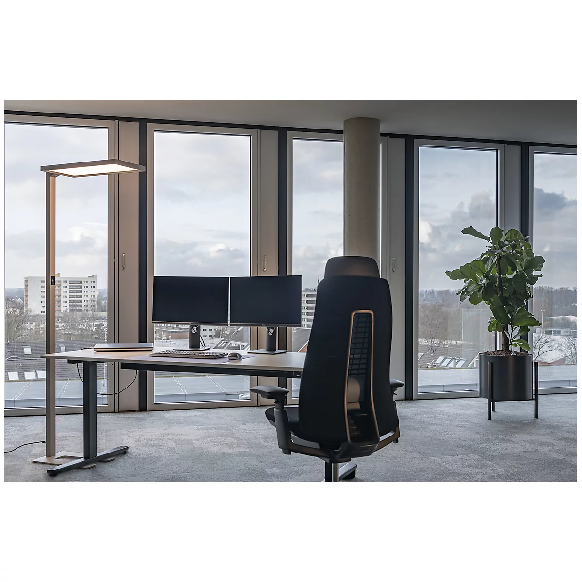 Bureau avec bureau, deux moniteurs, une chaise, une plante et une lampe devant des fenêtres panoramiques.