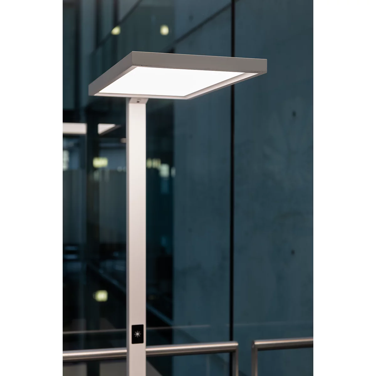 Lampadaire moderne avec un abat-jour carré et lumineux. Mât argenté, fond gris.