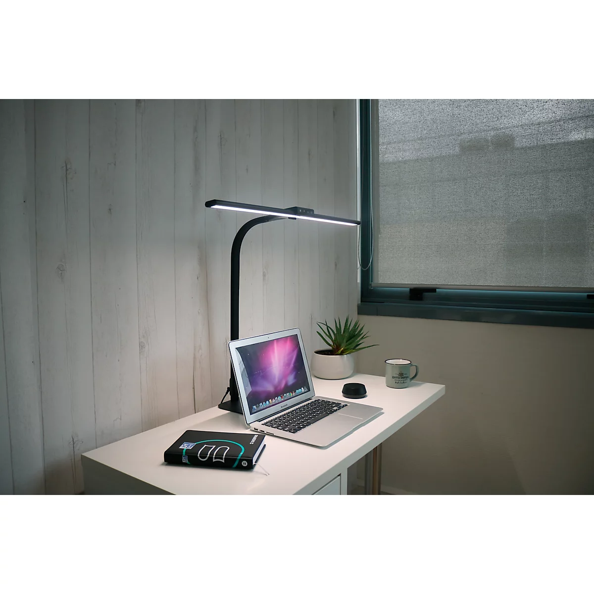 Une lampe de bureau noire éclaire un bureau. Sur la table, il y a un ordinateur portable, une plante, une tasse et une batterie externe.