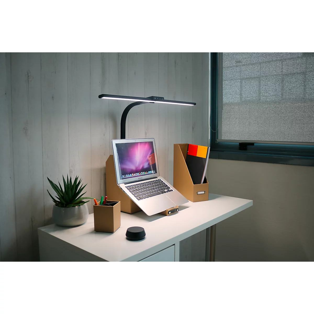 Bureau avec ordinateur portable, lampe et plante. Une lampe de lecture noire éclaire le cahier. À droite une fenêtre, à gauche une plante en pot.