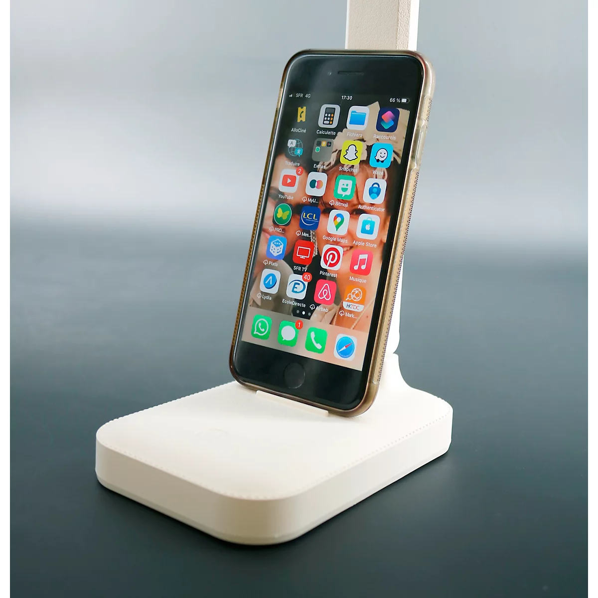 Smartphone sur support blanc. L'écran affiche des icônes d'applications. Le fond est gris.