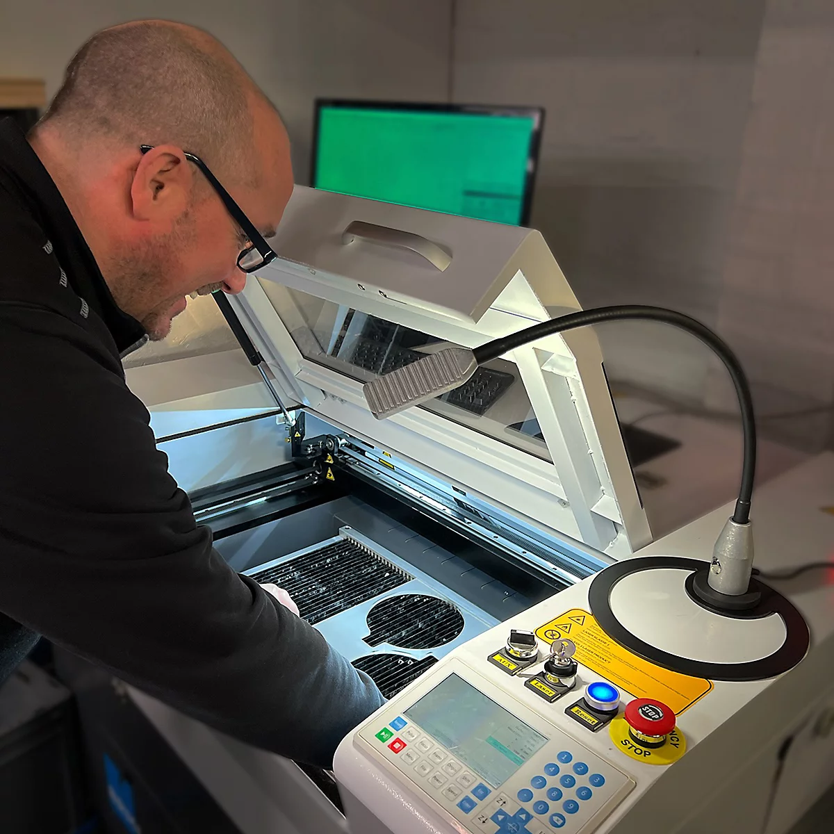 Un homme en pull noir travaille sur une machine de gravure laser. Il se penche sur la machine, éclairée par une lampe, et semble vérifier une pièce.