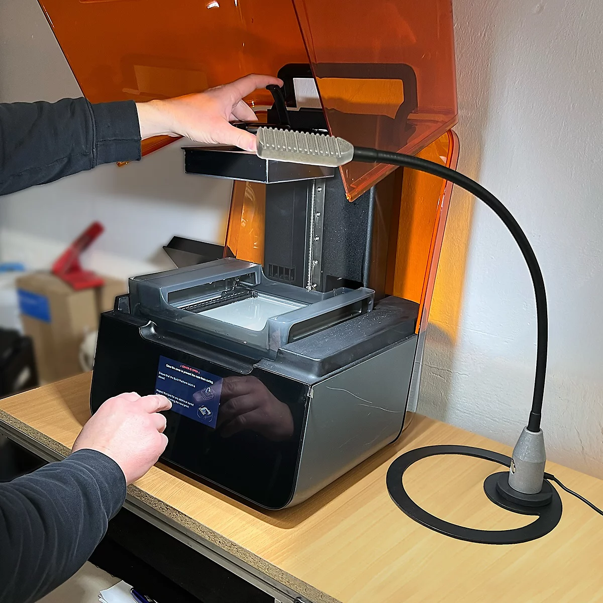 Une imprimante 3D avec un couvercle orange. Une main actionne l'écran tactile, une autre ouvre le capot supérieur. Une lampe flexible éclaire l'imprimante.