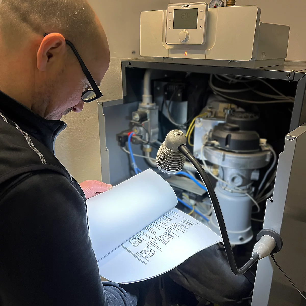 Un homme portant des lunettes inspecte un radiateur, lisant un manuel d'instructions sous une lampe.