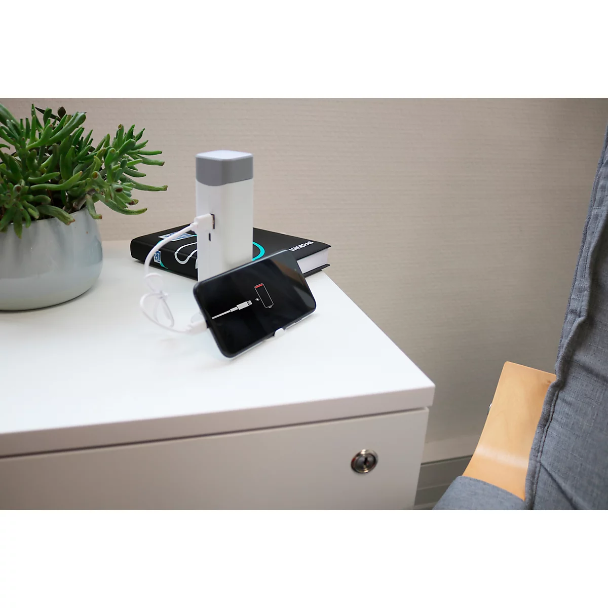 Un smartphone se charge sur une table de chevet blanche. A côté, un chargeur, un livre et une plante. En arrière-plan : une chaise.