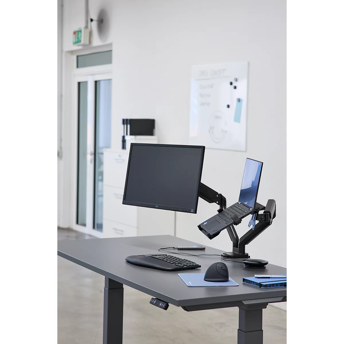 Schreibtisch mit Monitor, Laptop und Peripheriegerät. Helle Büroumgebung. Der Monitor und der Laptop sind auf Halterungen befestigt.
