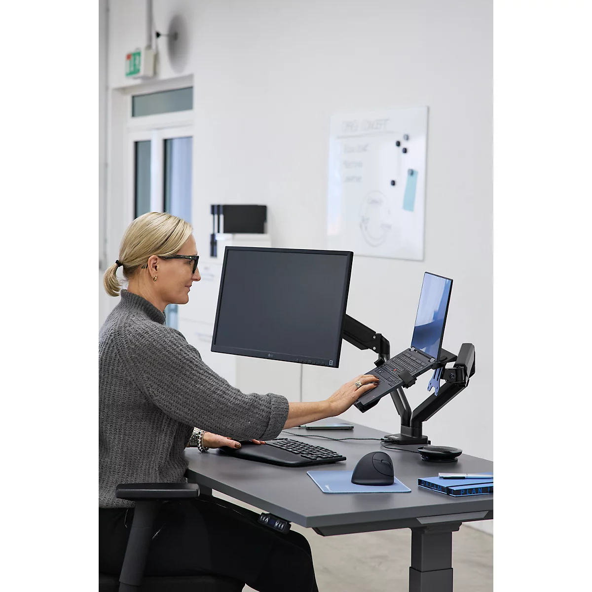 Eine Frau arbeitet an einem höhenverstellbaren Schreibtisch mit Monitor, Laptop, Tastatur und Maus. Sie benutzt den Laptop.