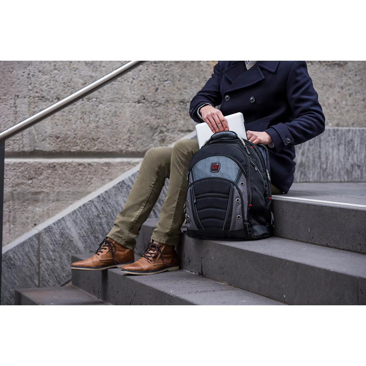 Ein Mann sitzt auf einer Treppe und verstaut einen Laptop in seinem Rucksack. Er trägt eine blaue Jacke und braune Schuhe.
