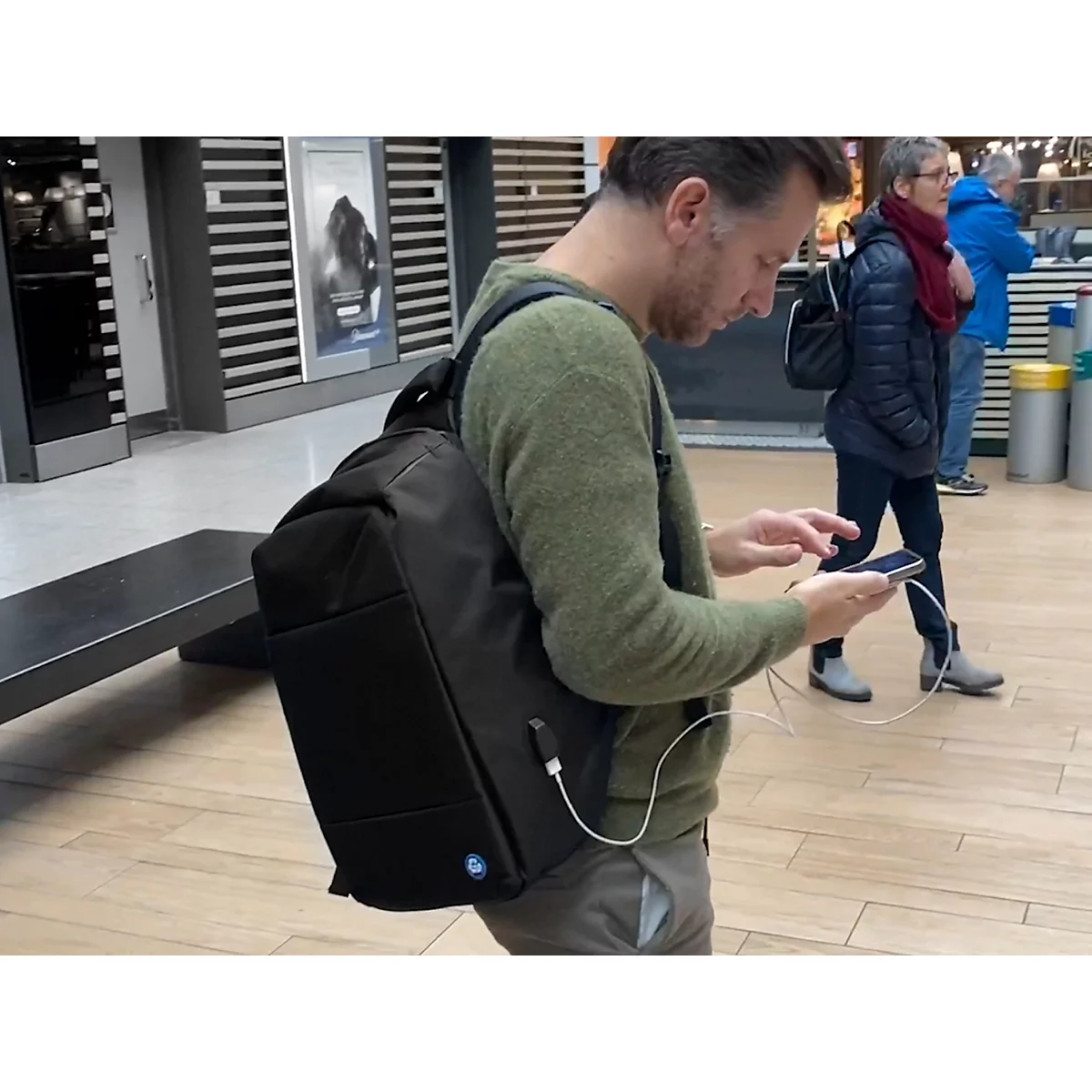 Een man met een rugzak aan de telefoon. Een witte oplaadkabel verbindt de telefoon met de rugzak. Achter hem staan andere mensen.