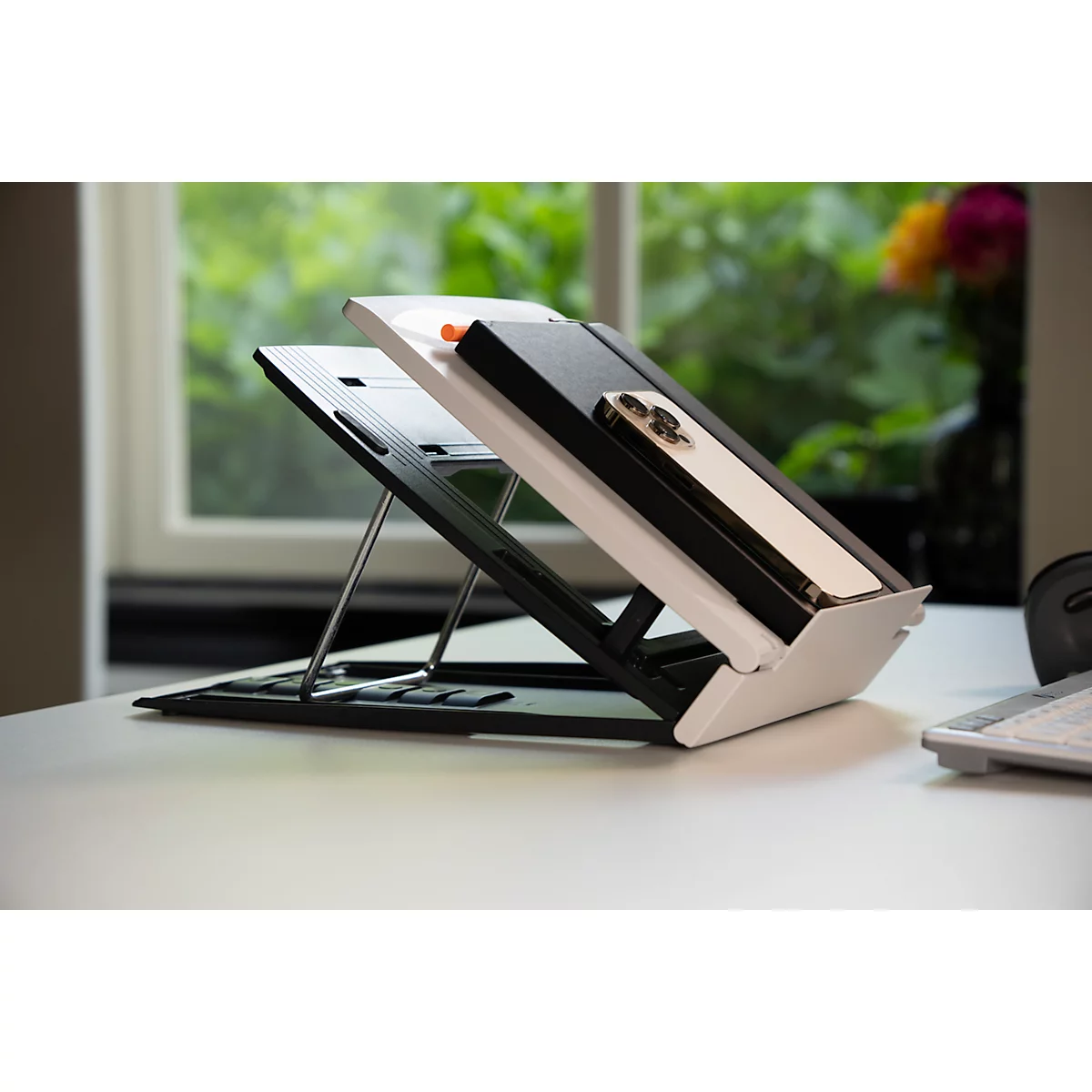 Zwarte en witte laptopstandaard met smartphone en notitieboek, op een bureau geplaatst, raam op de achtergrond.