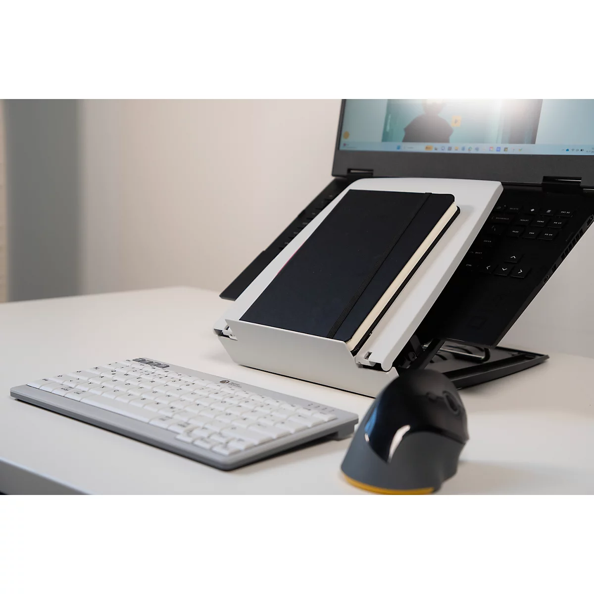 Laptop met notitieboek op standaard, toetsenbord en muis op witte tafel.