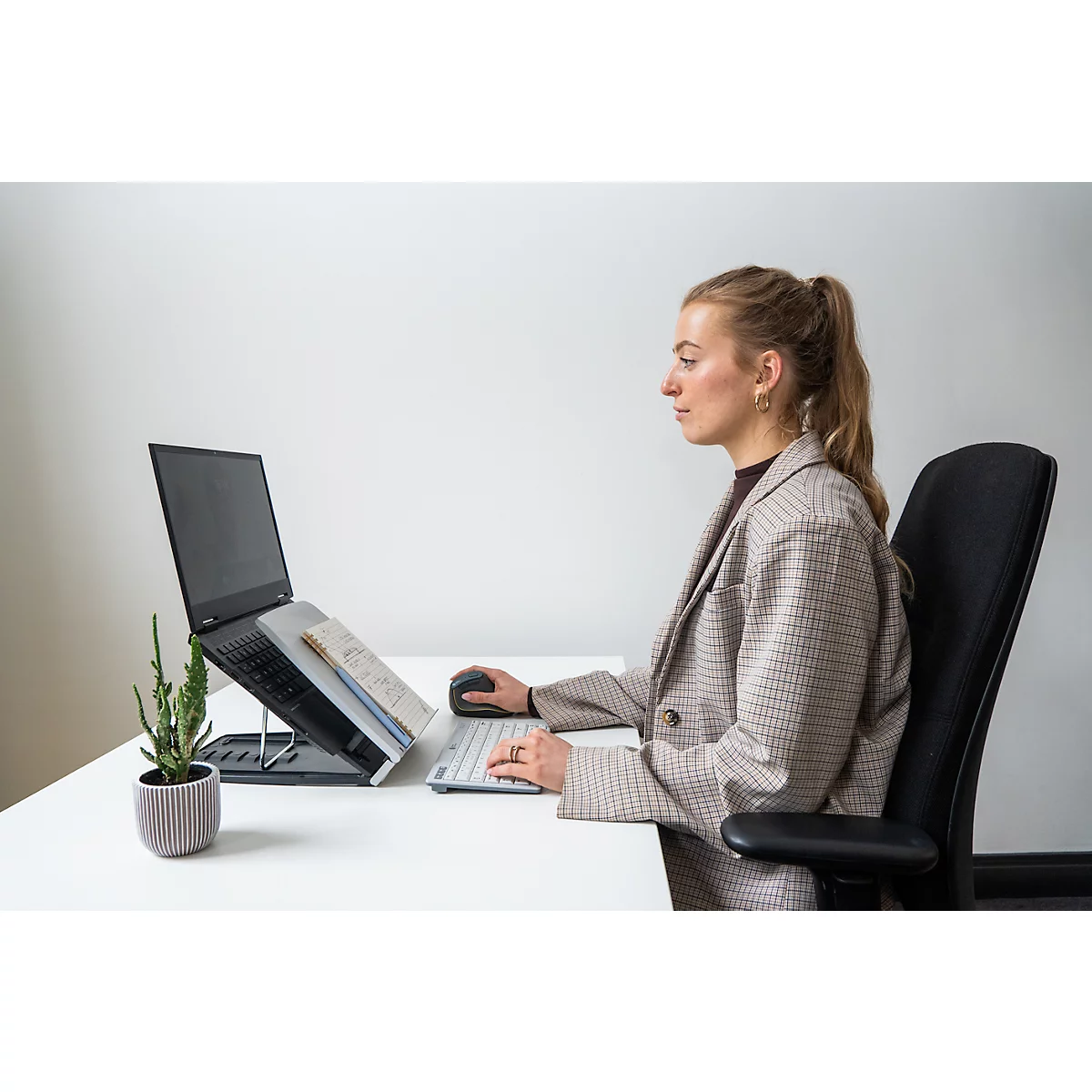 Een vrouw zit aan een bureau met een laptop, muis en toetsenbord. Ze draagt een blazer, haar haar is vastgebonden in een paardenstaart.