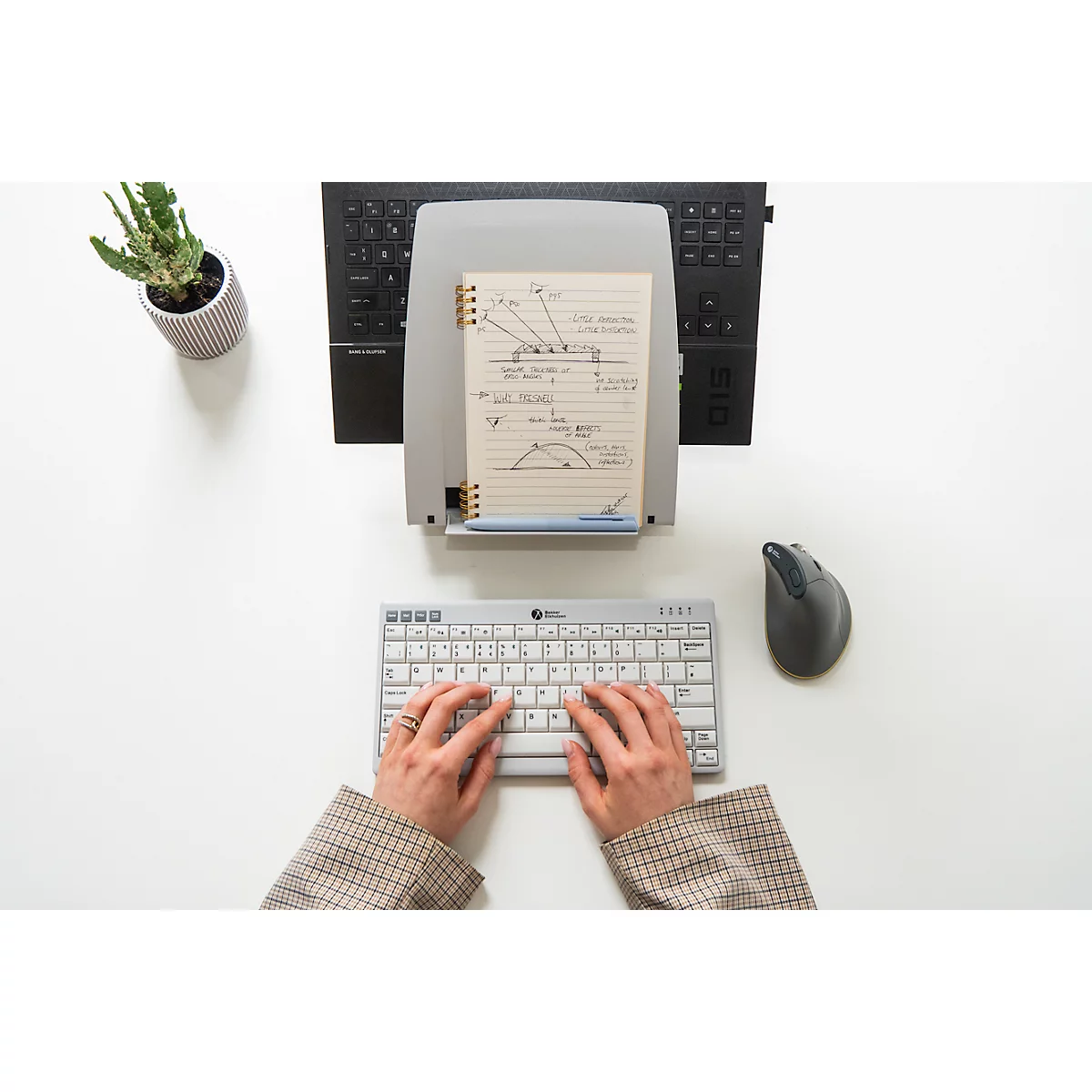 Bovenaanzicht van een persoon die op een toetsenbord typt. Daarnaast een muis, notitieblok en een plant.