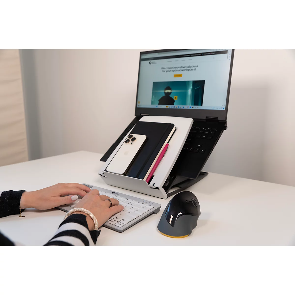 Vrouw tikt op een toetsenbord. Op de tafel: laptop met website, smartphone, notitieboek, muis.