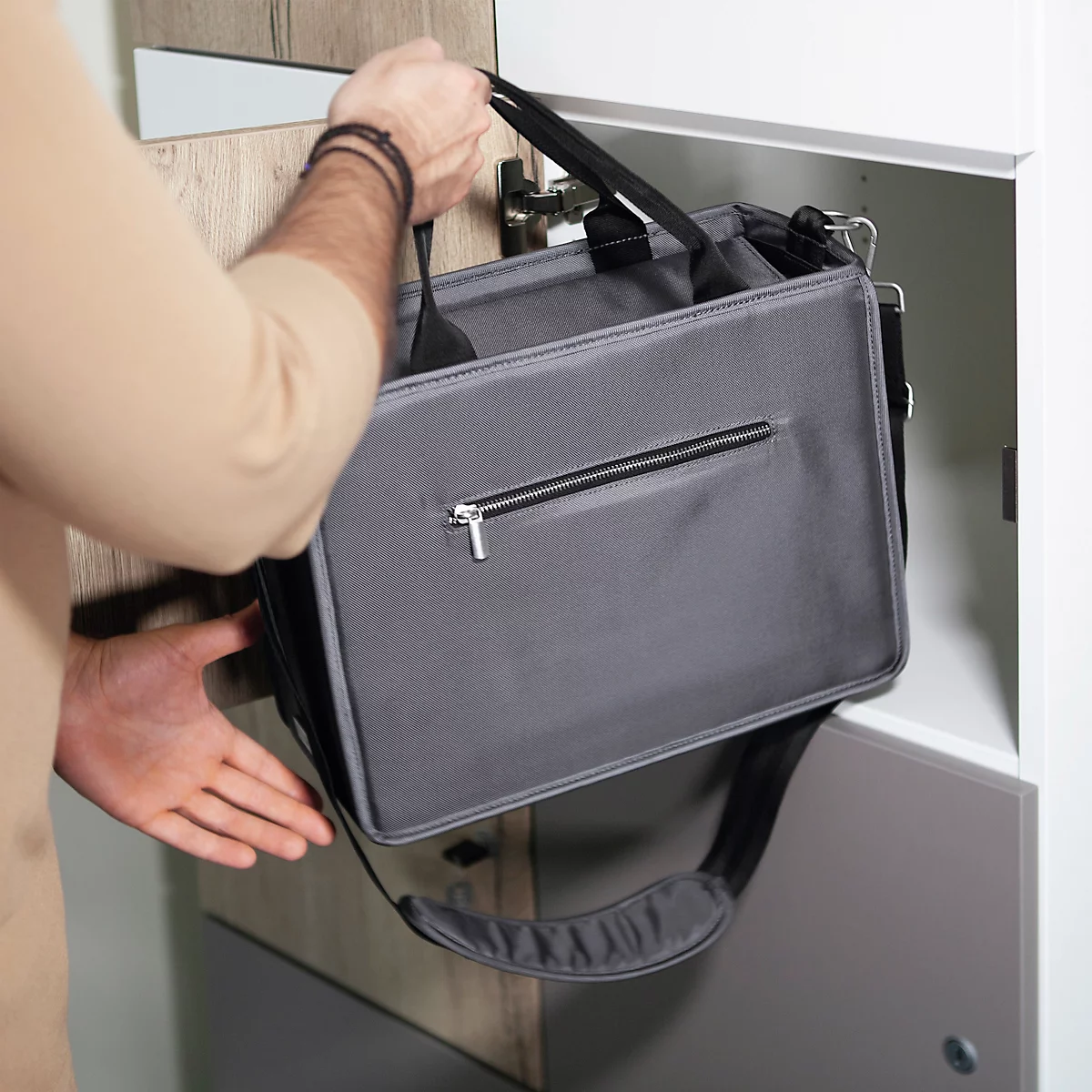 Person hält graue Laptoptasche. Rechter Arm liegt im Schrank. Schwarz-grauer Schultergurt.