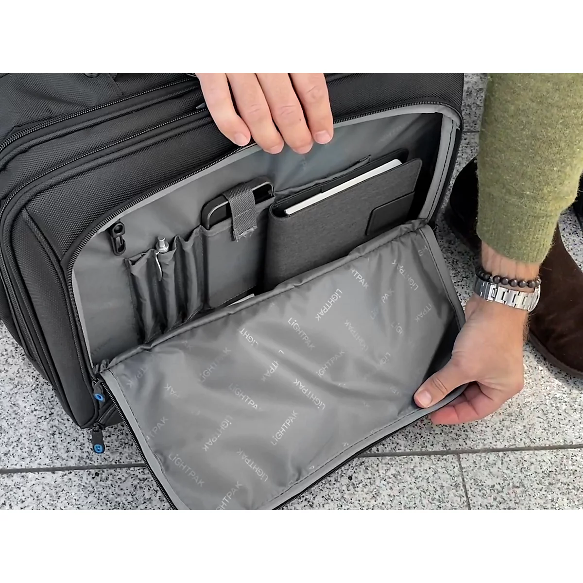 Person öffnet eine schwarze, offene Handgepäcktasche. Innenfächer für Laptop, Handy und Stifte sind zu sehen.