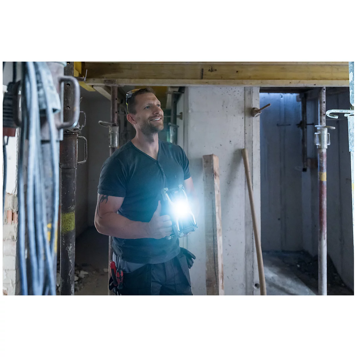Ein Handwerker lächelt und hält eine Taschenlampe, während er in einem Baustellenbereich steht. Licht strahlt von der Lampe.
