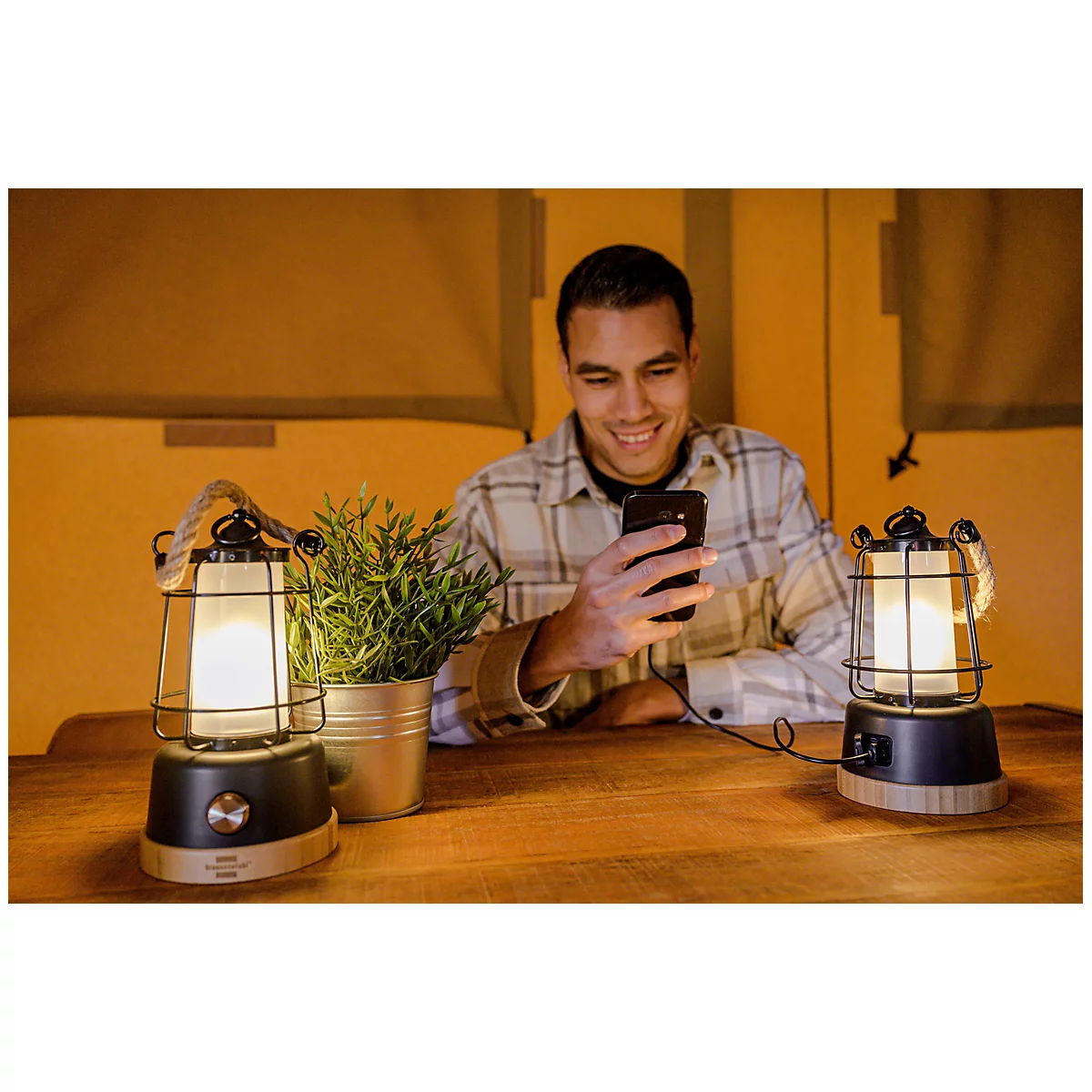 Ein junger Mann in einem karierten Hemd sitzt an einem Tisch, lächelt und schaut auf sein Smartphone, umgeben von zwei Laternen und einer kleinen Pflanze.