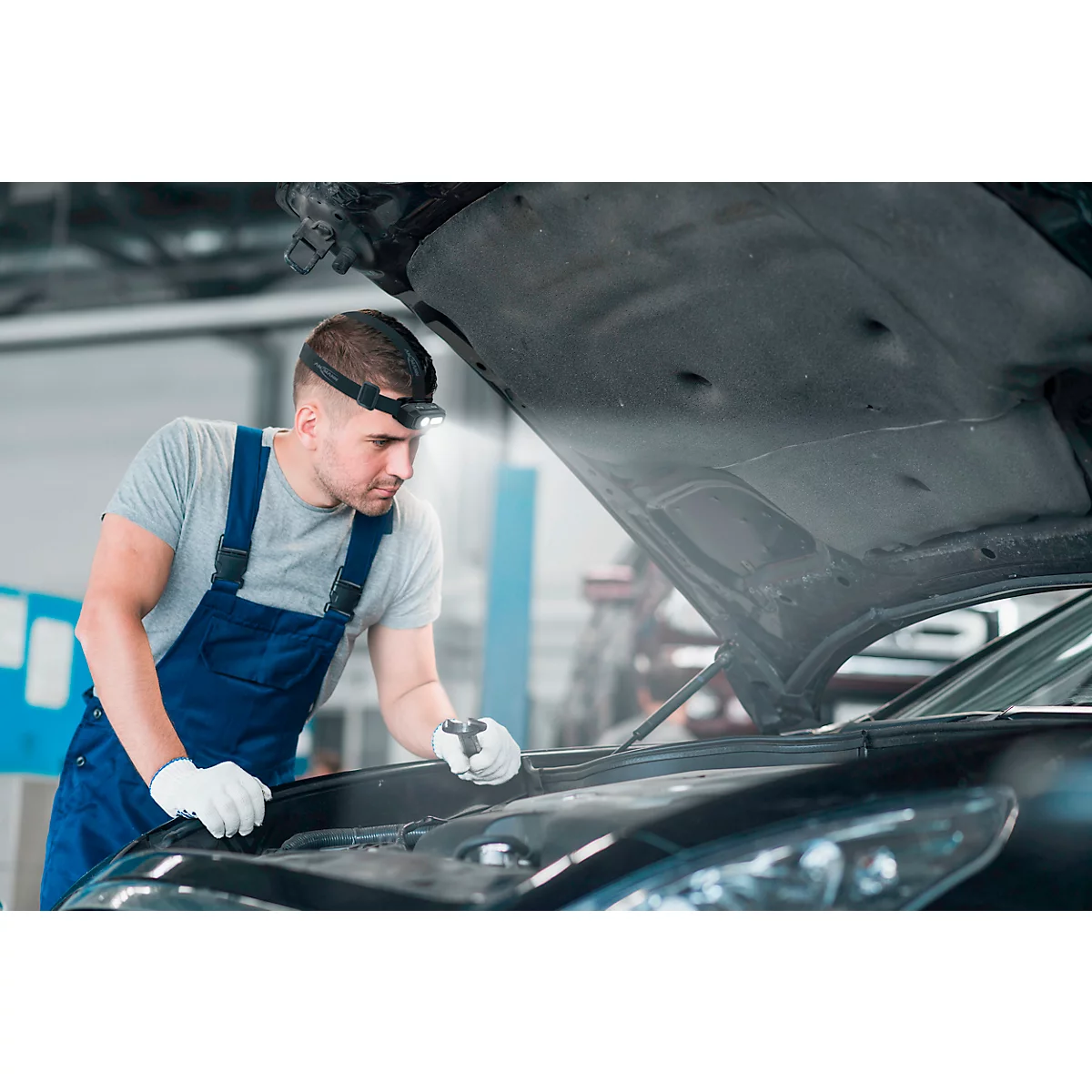 A mechanic with a headlamp looks under the hood of a car, wearing white work gloves.