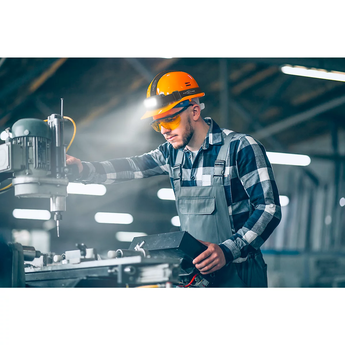 Man in work clothes at a machine. He wears an orange helmet with light, safety glasses. Beam of light.