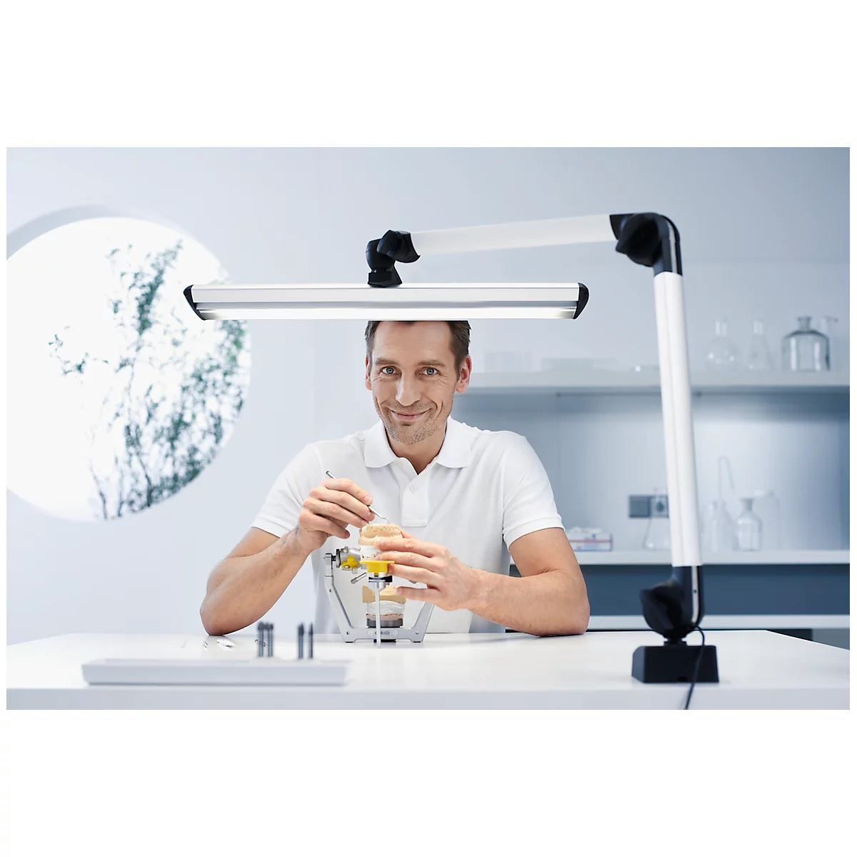 Dentist with a dental model. He smiles at the camera. Above him, a desk lamp.