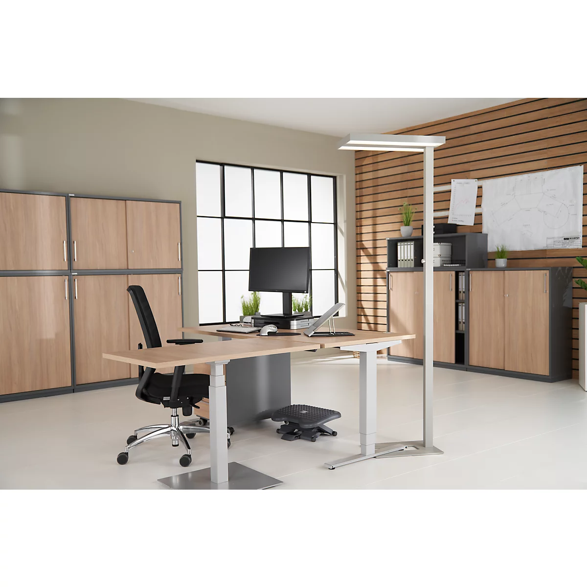 Heller, moderner Bürotisch mit Stuhl, Monitor, Laptop und Stehleuchte. Im Hintergrund Schränke und ein Fenster.