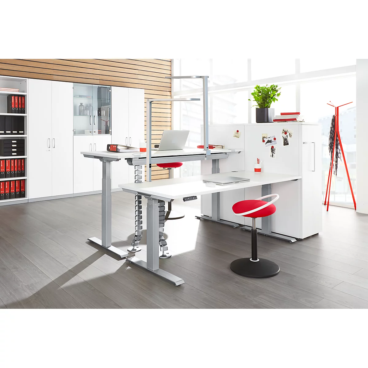 A modern office with height-adjustable desks and a red chair. White cabinets, plants, and a red coat rack complement the image.