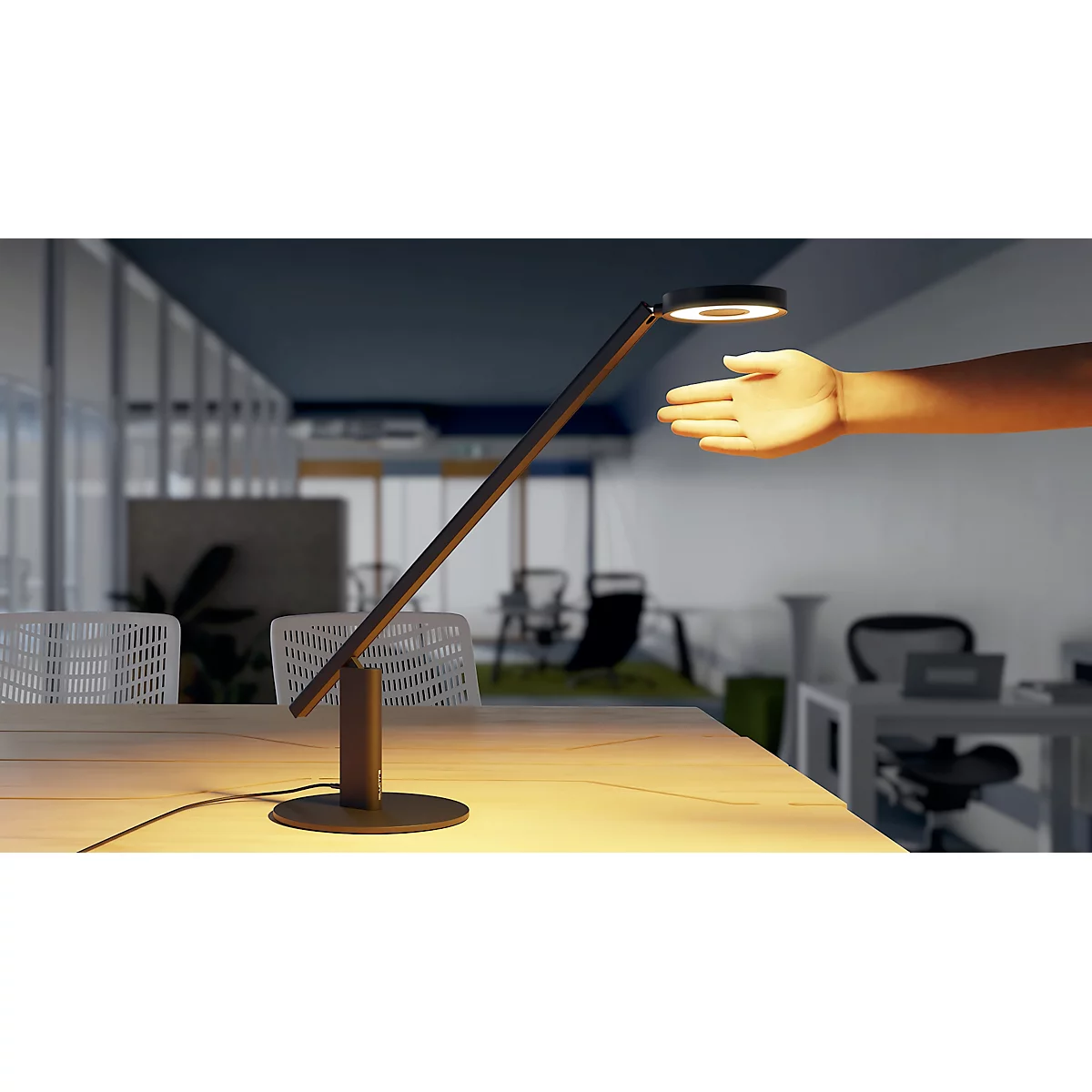 A stylish desk lamp with an arm, standing on a table. A hand is approaching the light. In the background an office.