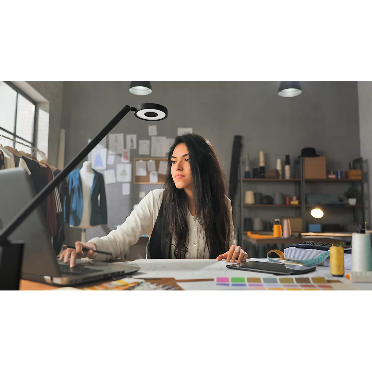 Designer working on laptop surrounded by fabric swatches, sketches, and lighting.