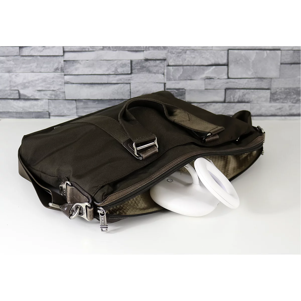 Braune Aktentasche, halb geöffnet. Eine weiße, moderne Schreibtischlampe liegt darin. Im Hintergrund eine graue Backsteinmauer.